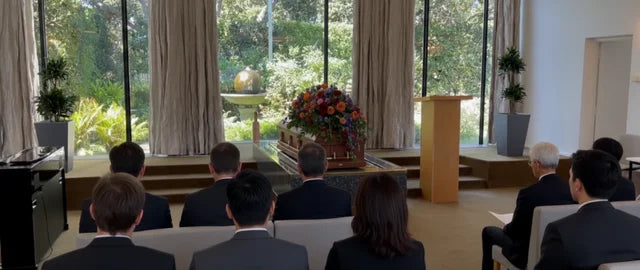 A traditional wooden casket covered with a large floral arrangement, placed in a bright, modern funeral chapel with natural light and guests seated for a service.