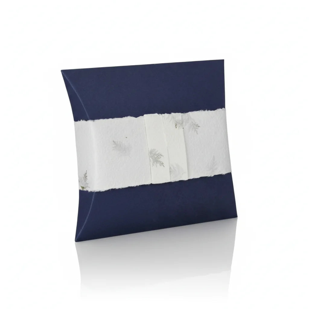 A high-quality product shot of a navy blue pillow-shaped urn with a decorative white band featuring silver leaf accents, presented on a clean white reflective surface with soft, even lighting