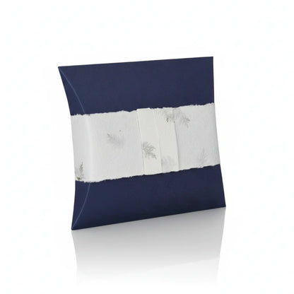 A high-quality product shot of a navy blue pillow-shaped urn with a decorative white band featuring silver leaf accents, presented on a clean white reflective surface with soft, even lighting