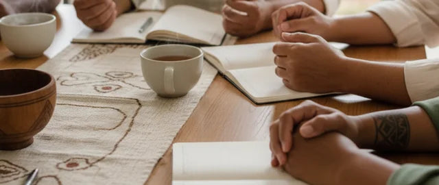 An overhead, close-up view of four sets of hands and arms (no faces visible) of a Cook Islands family gathered around a wooden table. They are wearing light, neutral-toned clothing (cream and soft green). In front of each person is an open notebook, suggesting a discussion. There are three cups of warm, steaming tea and a wooden bowl on a table runner with subtle, woven Pasifika-inspired patterns. The scene is softly lit, conveying calm, togetherness, and cultural support.