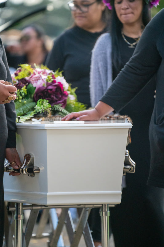 Close-up of a white casket/coffin being carried by family or funeral attendees during a service. A person's hand rests gently on the lid next to a vibrant bouquet of purple and green flowers. People dressed in black stand in the background
