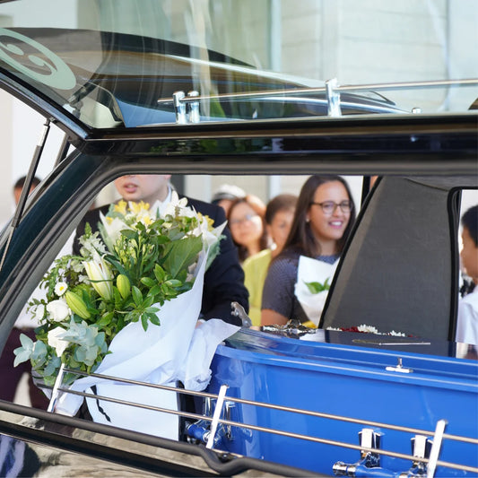 Close-up of a blue casket inside a hearse with a large bouquet of white and yellow flowers beside it. Several smiling mourners and family members are visible in the reflection and background