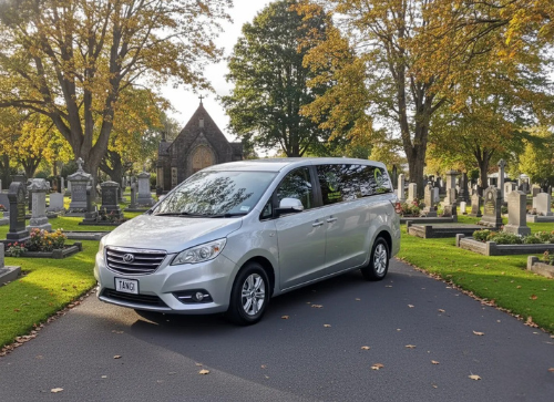 Silver coach hearse parked on a road with a cemetery in the background